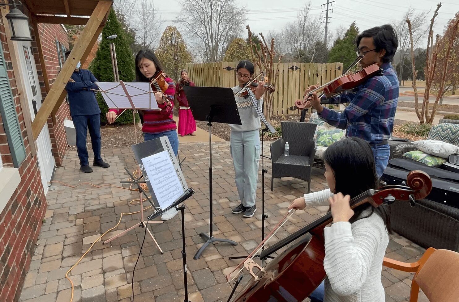 Group of people playing string instruments outdoors on a paved area with a wooden fence and trees in background.