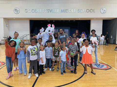 Group of children and adults in a gymnasium with a mascot costume of a white bunny with pink ears, in front of a stage. 20 words.