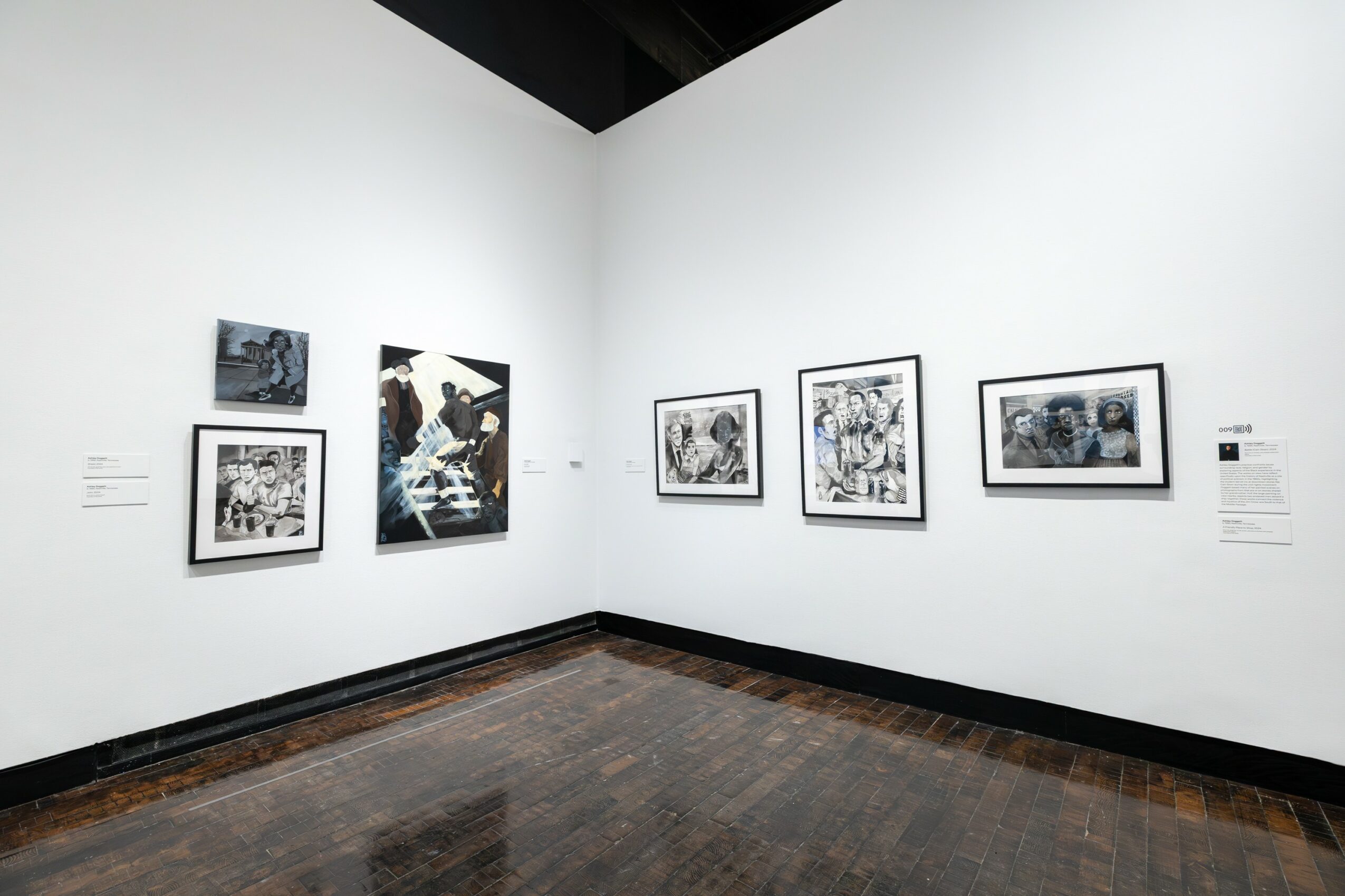 Gallery wall with framed black-and-white and color artworks, white walls, wooden floor, and labels beside each piece.