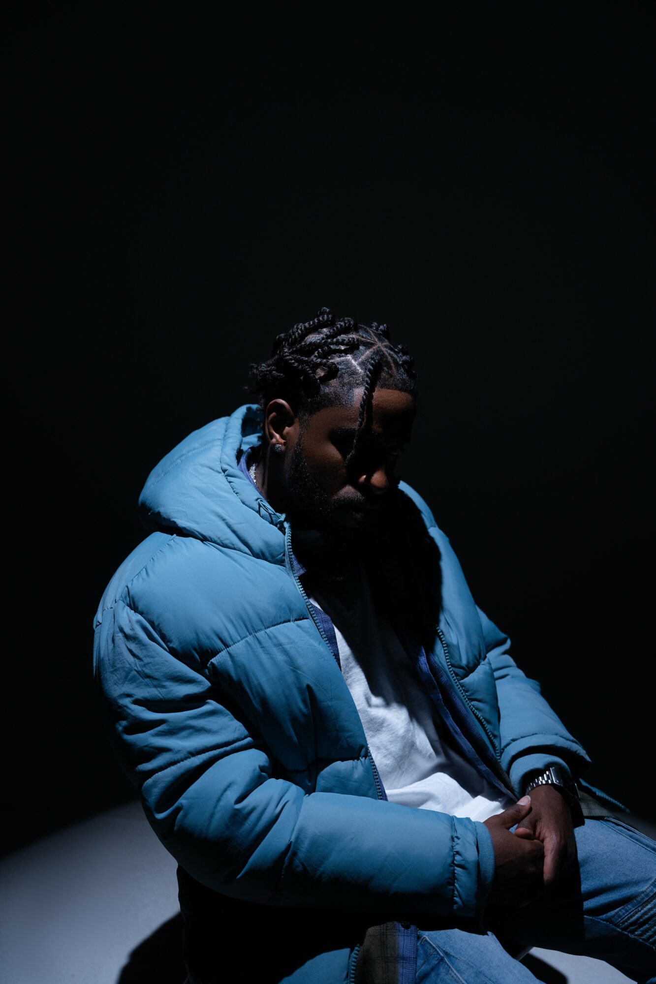 Person with braided hair wearing a blue puffer jacket, head bowed, in a dark setting.