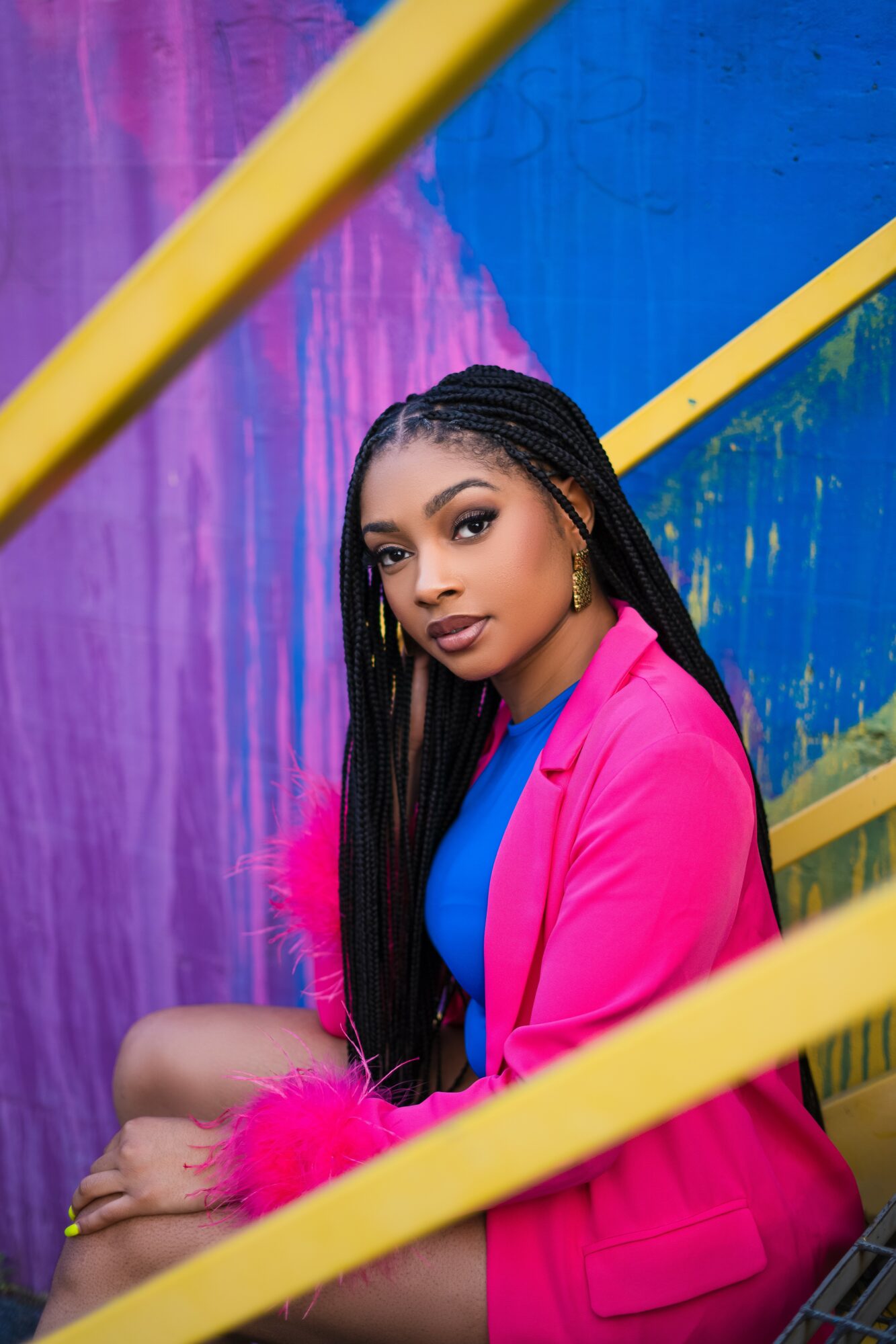 Young woman with long braided hair wearing a pink blazer and blue top, sitting against a colorful abstract background.