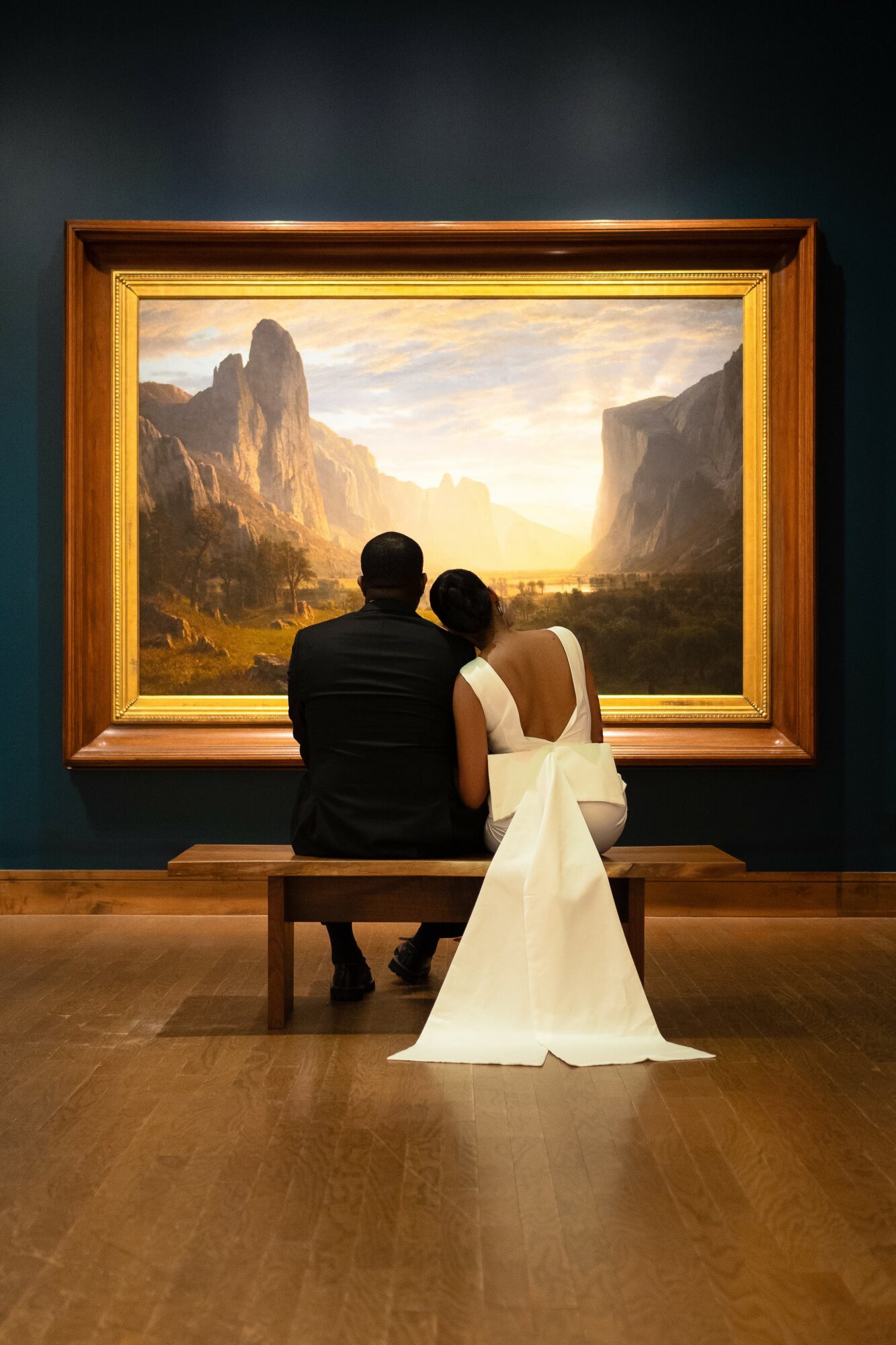 Two people sit on a bench viewing a landscape painting in a gallery, with mountains and a sunset sky.