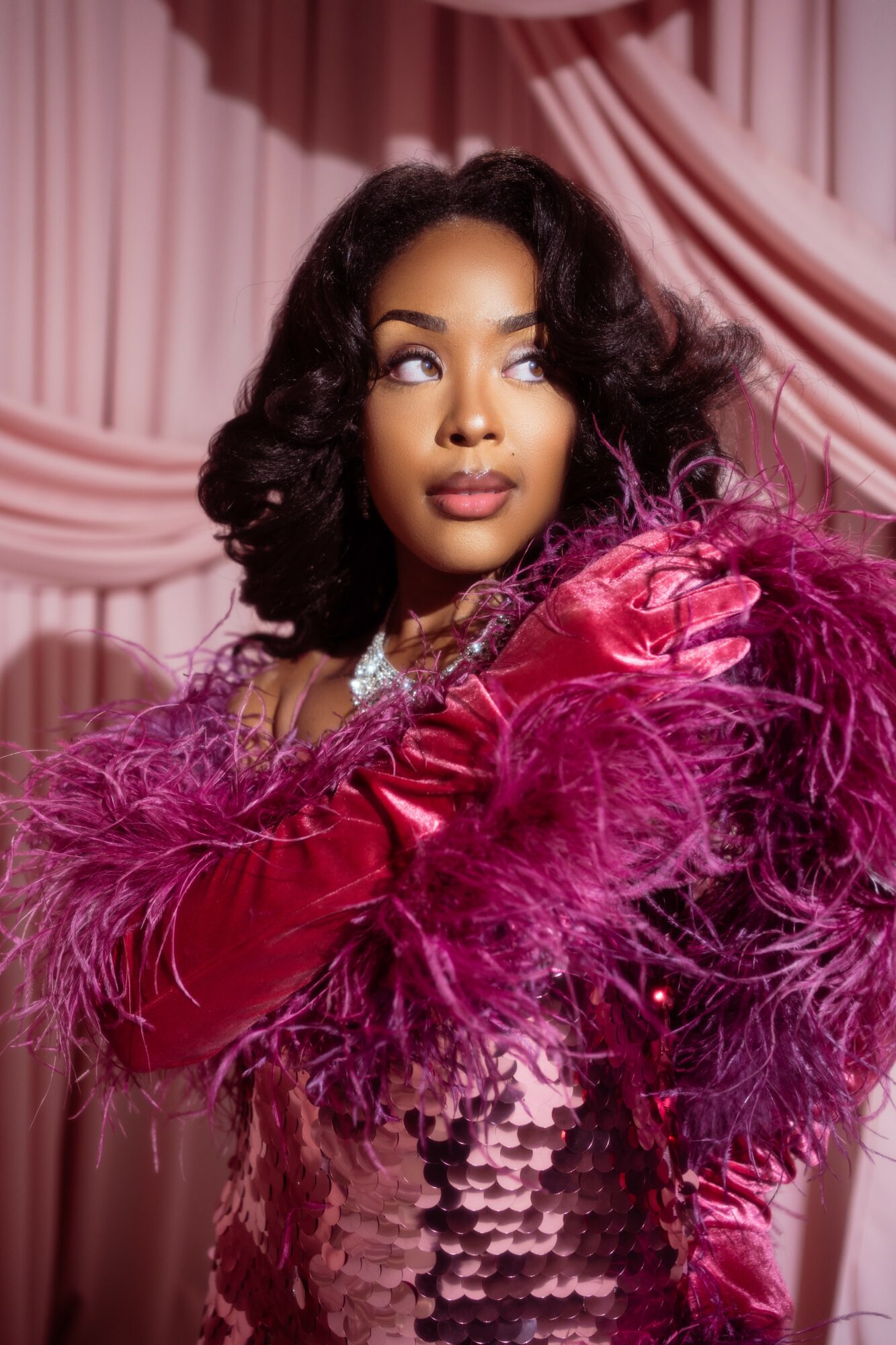 Woman with curly hair wearing a pink feathered garment, standing in front of pink draped curtains.