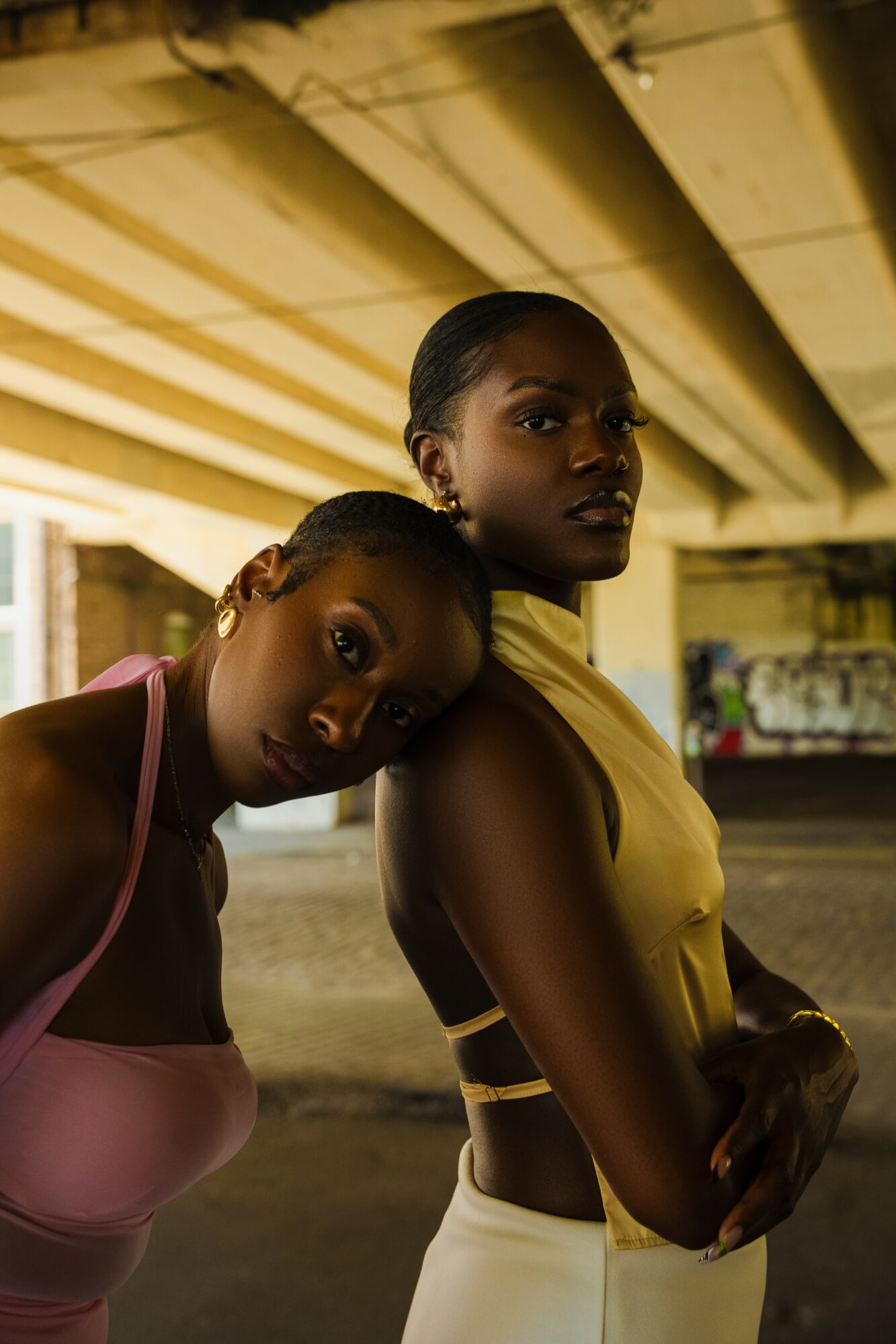 Two women under a bridge, one leaning on the other's shoulder, both looking at the camera, in a dimly lit urban setting.