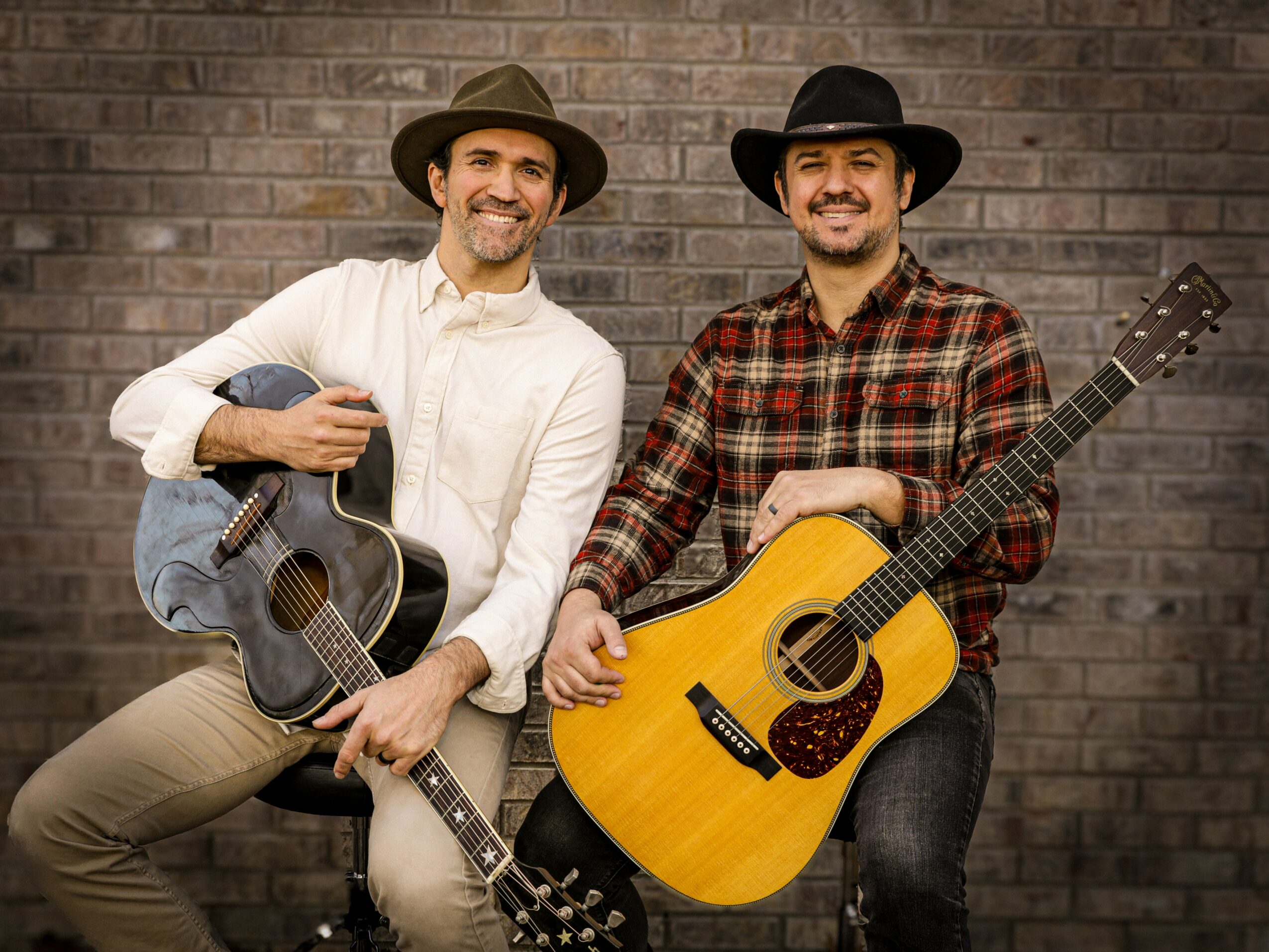 Two men with guitars standing against a brick wall, smiling, wearing hats and casual clothing.