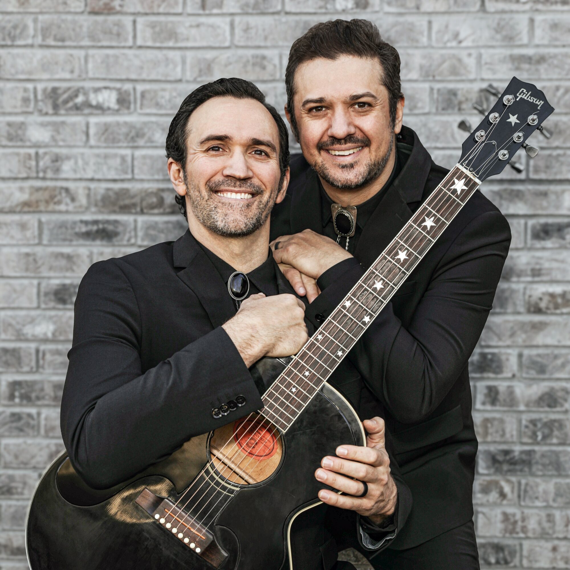 Two men smiling, one holding a guitar, standing against a brick wall, dressed in black suits.