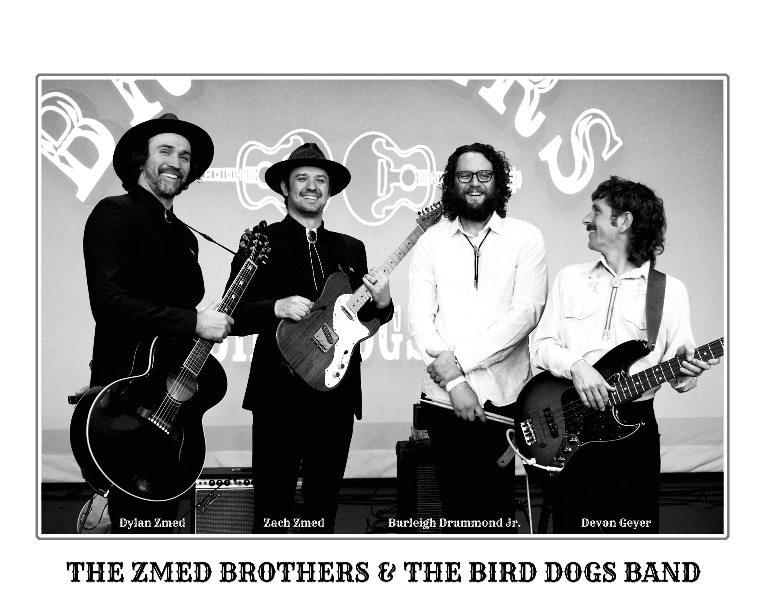 Four men holding guitars and smiling, standing in front of a backdrop with band names, black and white photo.