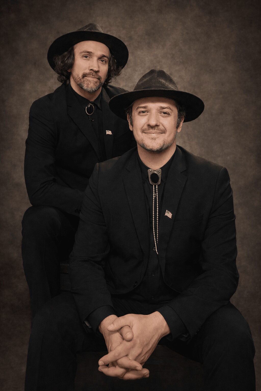 Two men in black suits and hats pose against a dark background, one sitting and one standing, both smiling.