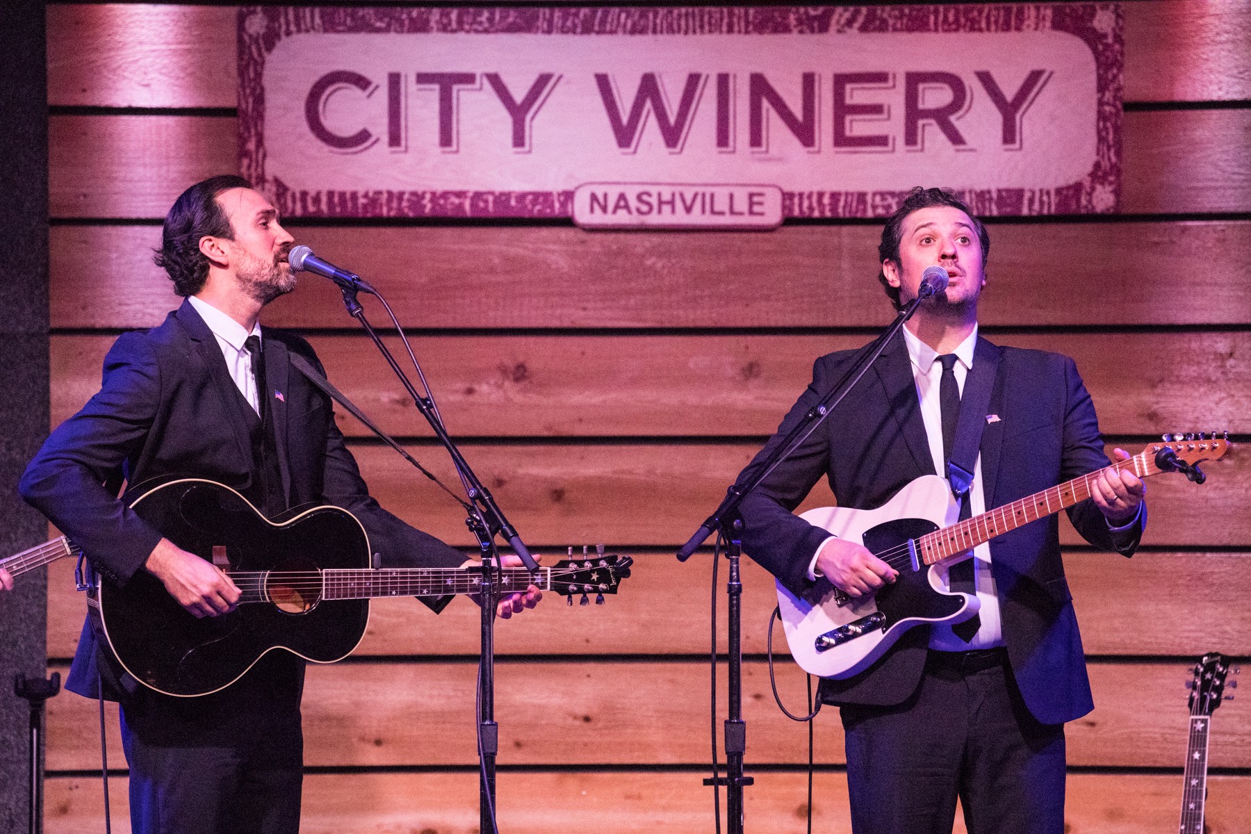 Two men in suits playing guitars and singing on stage with a wooden backdrop and a sign reading 'City Winery Nashville'.
