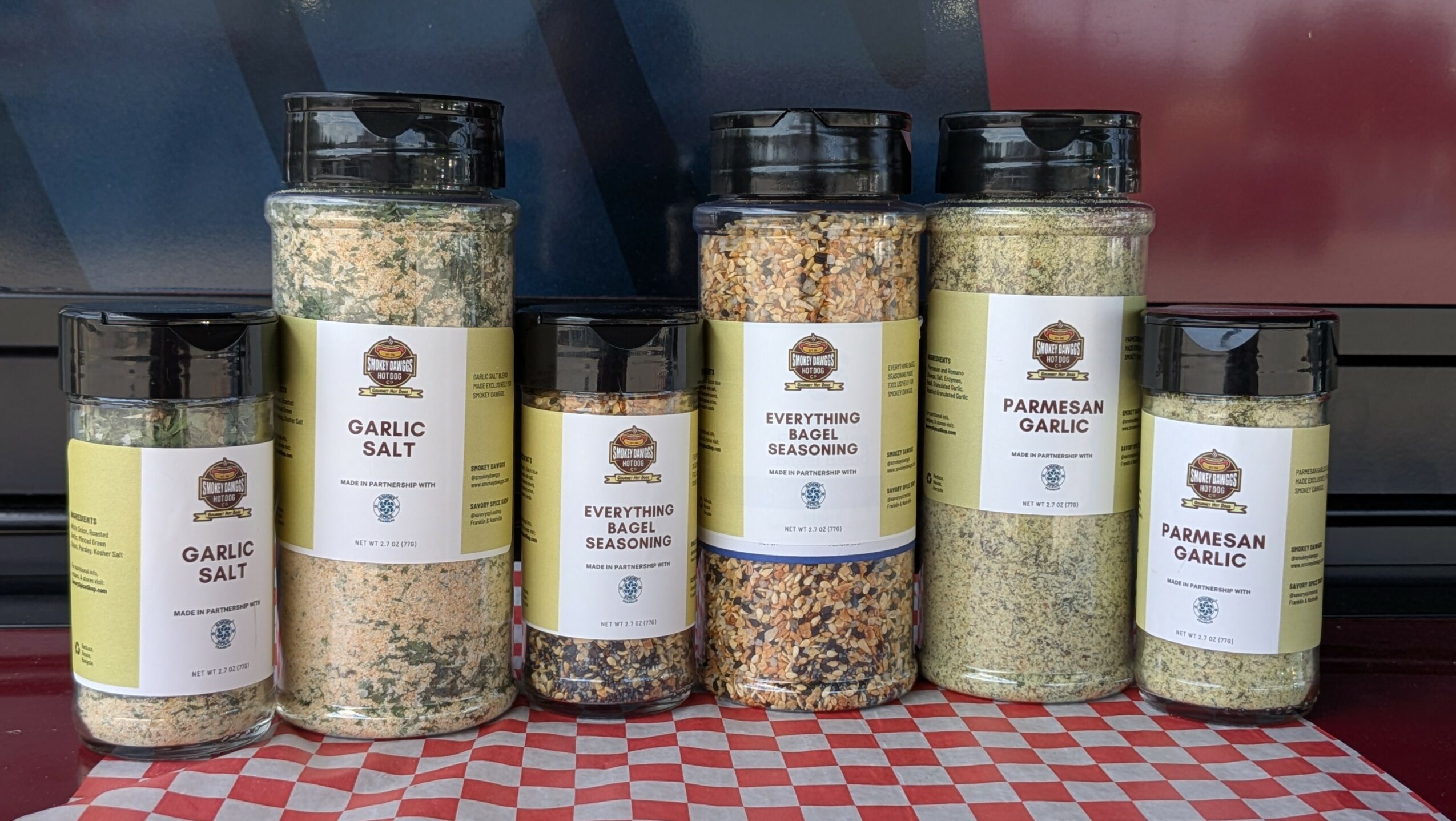 Six spice jars with black lids and white labels on a checkered surface, containing garlic salt, seasoning, and Parmesan garlic.