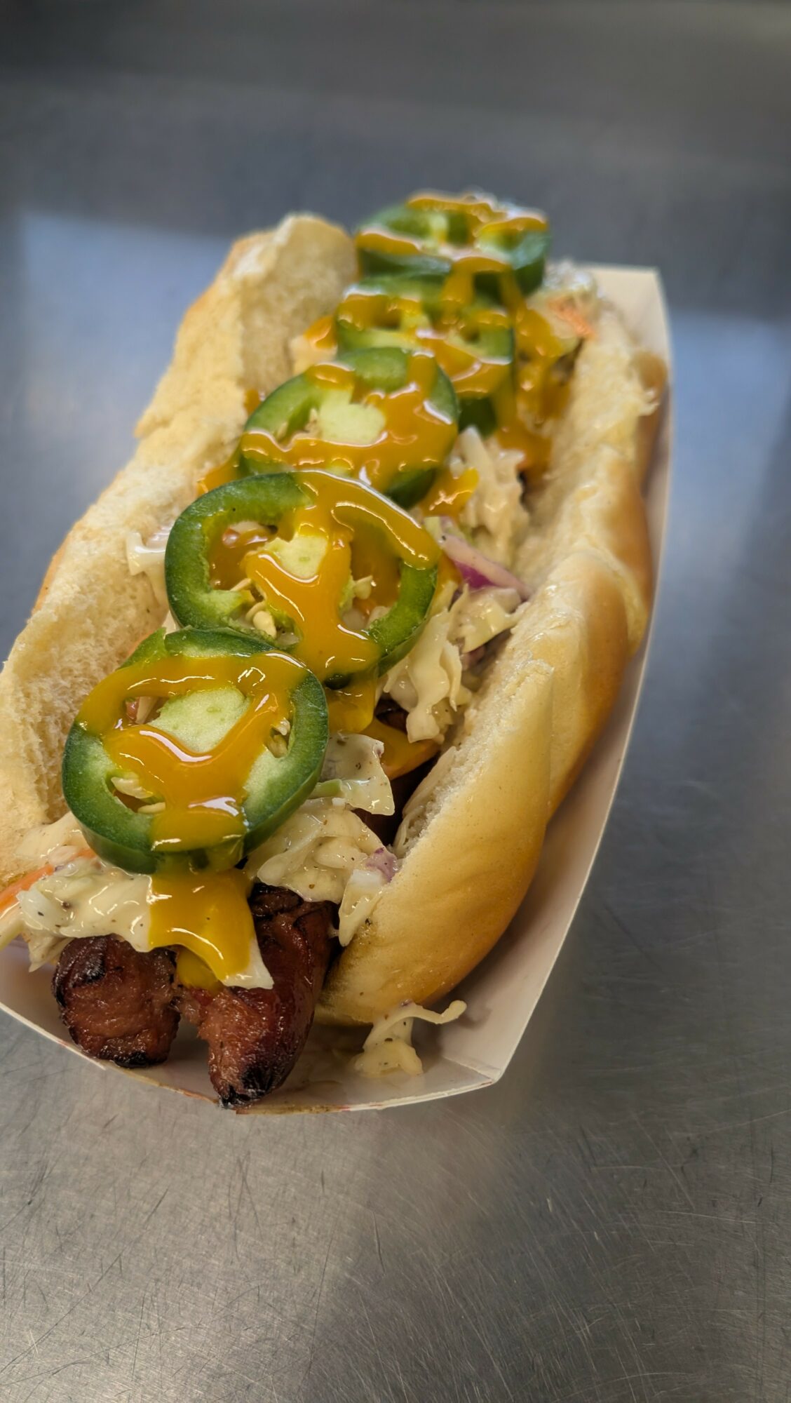 Hot dog with mustard, jalapeños, shredded cheese, and fried onion on a bun, served in a paper tray.