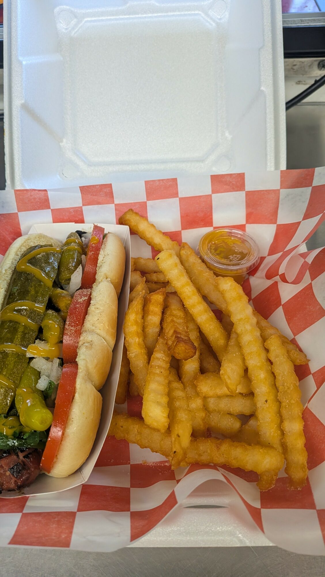 Hot dog with pickles and mustard, crinkle-cut fries, and a small container of dipping sauce on red and white checkered paper.