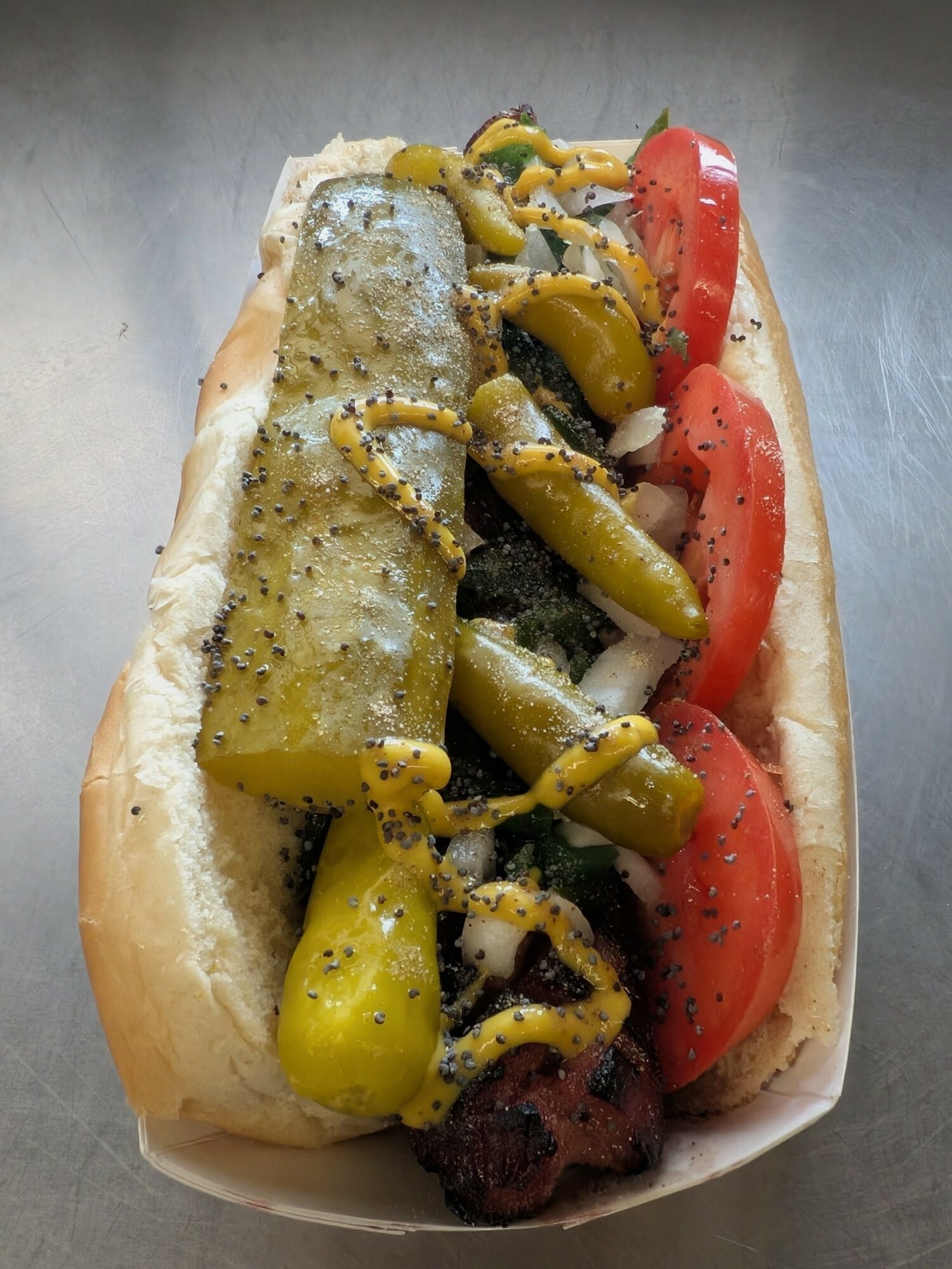 Hot dog bun filled with sliced tomatoes, pickles, onions, and black pepper, served in a paper tray.