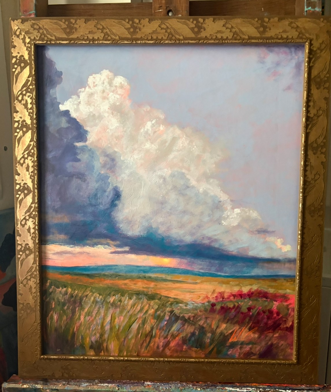 Landscape painting of a sky with clouds over fields, framed in an ornate gold frame.