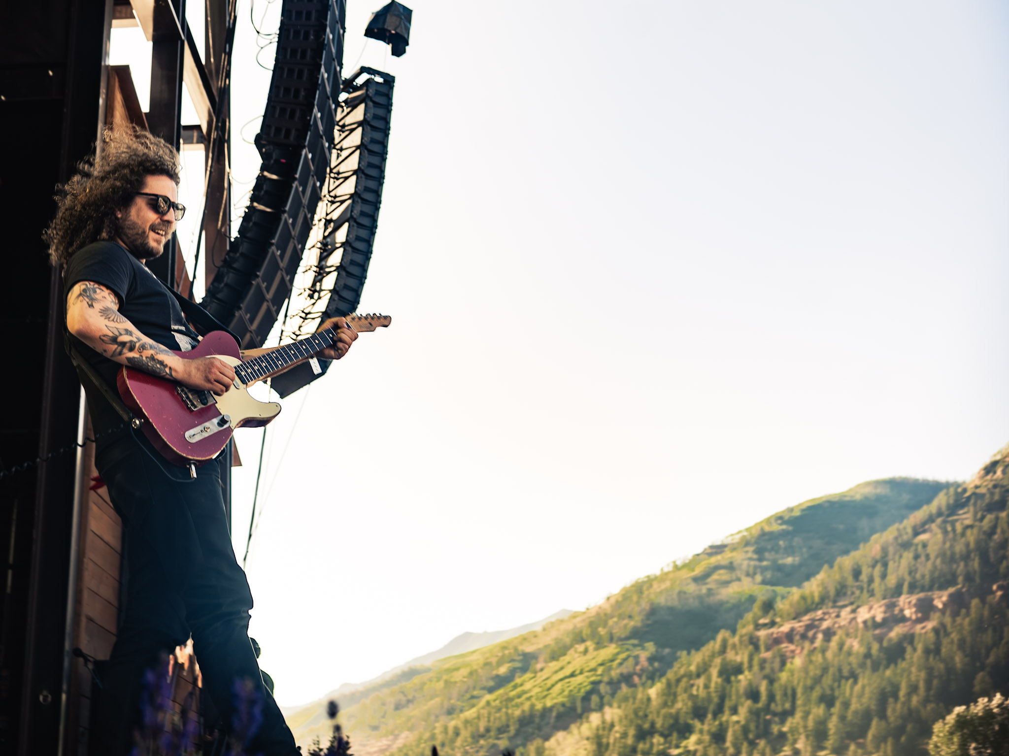 Person with curly hair and sunglasses playing a pink guitar outdoors, mountains in the background.