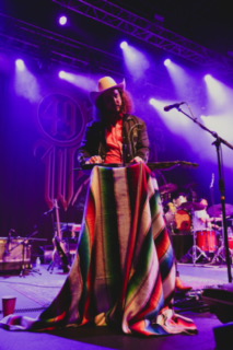 Person wearing a hat and colorful blanket on stage with musical instruments and purple lighting.