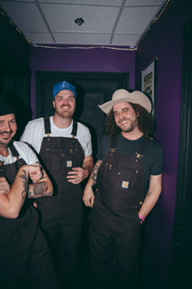 Three people in black overalls and hats, smiling in a purple-walled room.