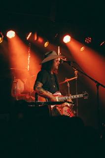 Musician wearing a hat playing guitar on stage with orange and yellow stage lights, smoke, and microphone.