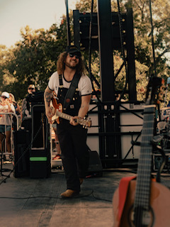 Musician with long hair, sunglasses, playing guitar outdoors during daytime, surrounded by trees and people, with equipment in background.
