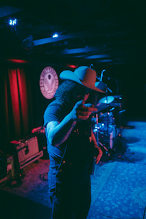 Person wearing a large hat performing on stage with drums and a clock in the background, dimly lit with red curtains.