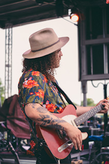 Person wearing a hat playing a pink guitar outdoors, with a structure and sky in the background.