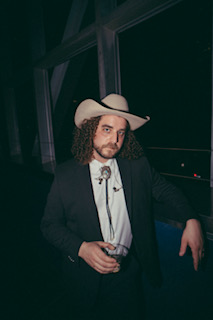 Man with curly hair and beard wearing a cowboy hat, black suit, and white shirt, holding a glass, standing indoors.