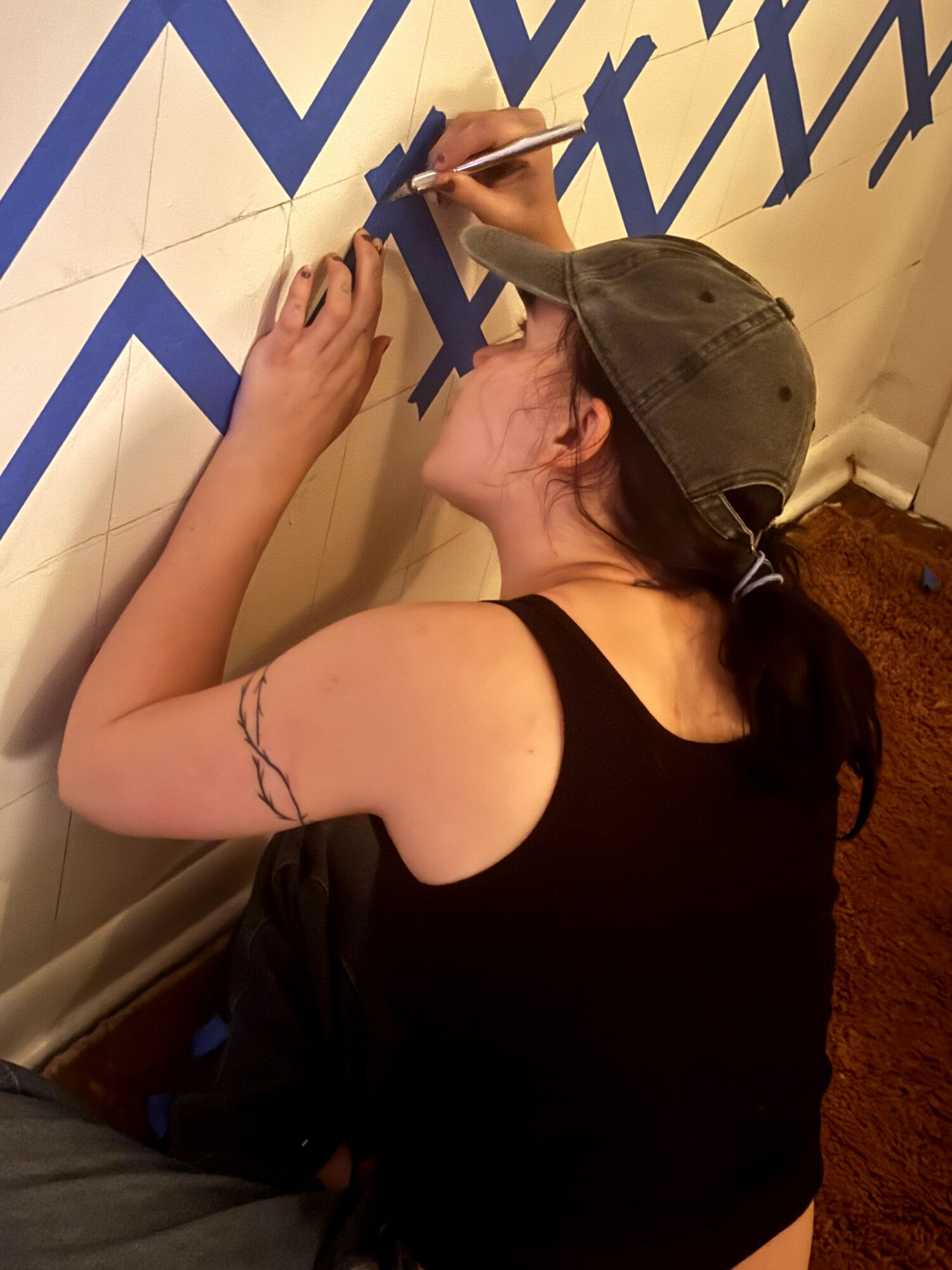 Person with a tattoo on arm, wearing a cap, writing on wall with geometric pattern, in a room with carpet.