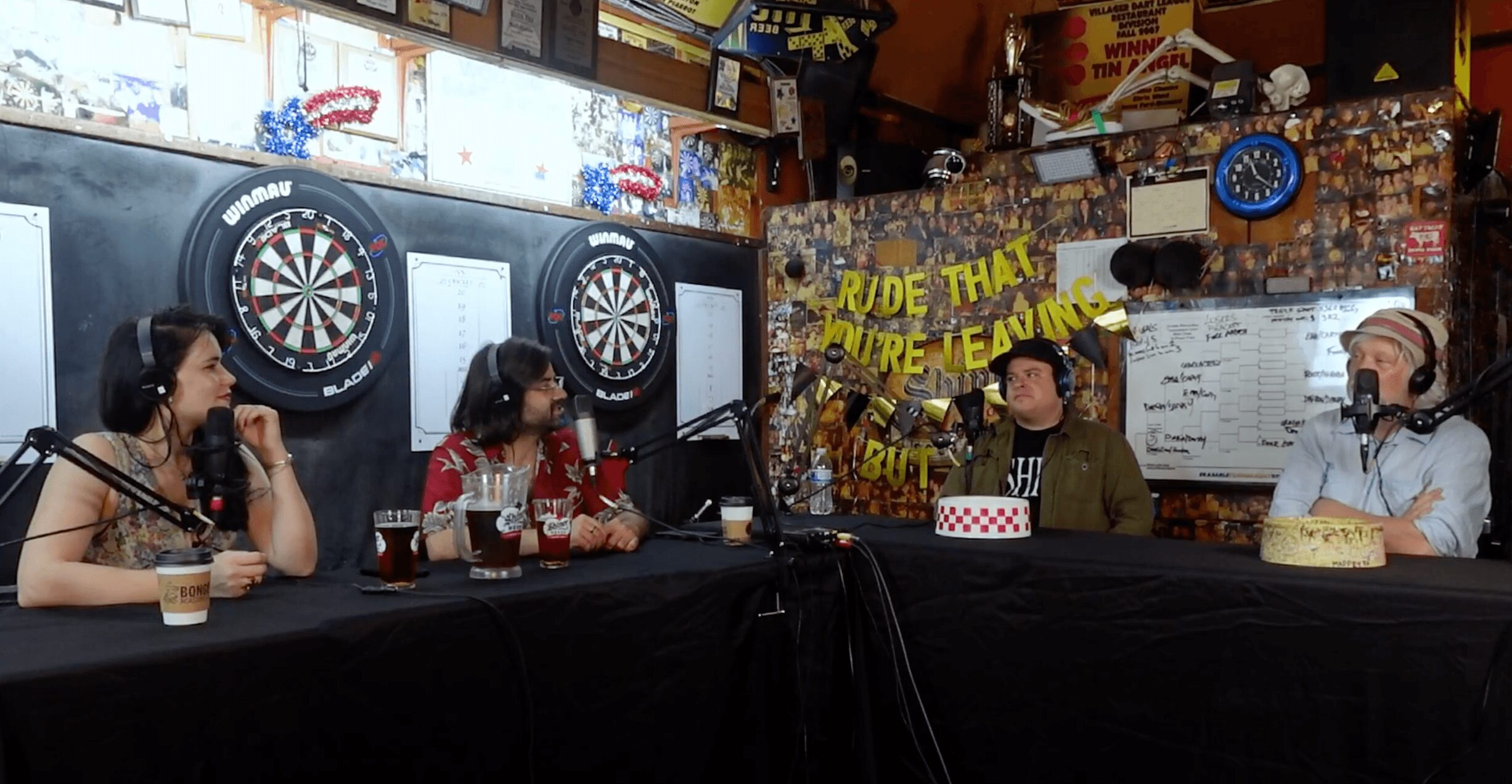 People sitting at a table in a bar with dartboards on the wall, some wearing headphones, and a yellow sign in the background.