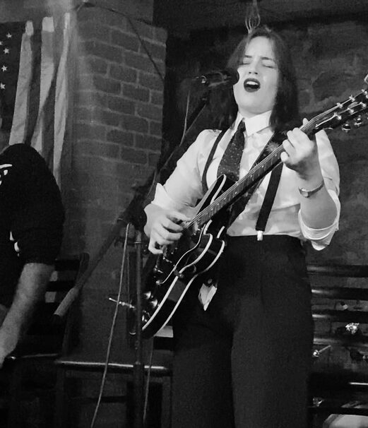 Woman singing and playing guitar on stage with brick wall background, microphone in front, eyes closed, expressive performance.