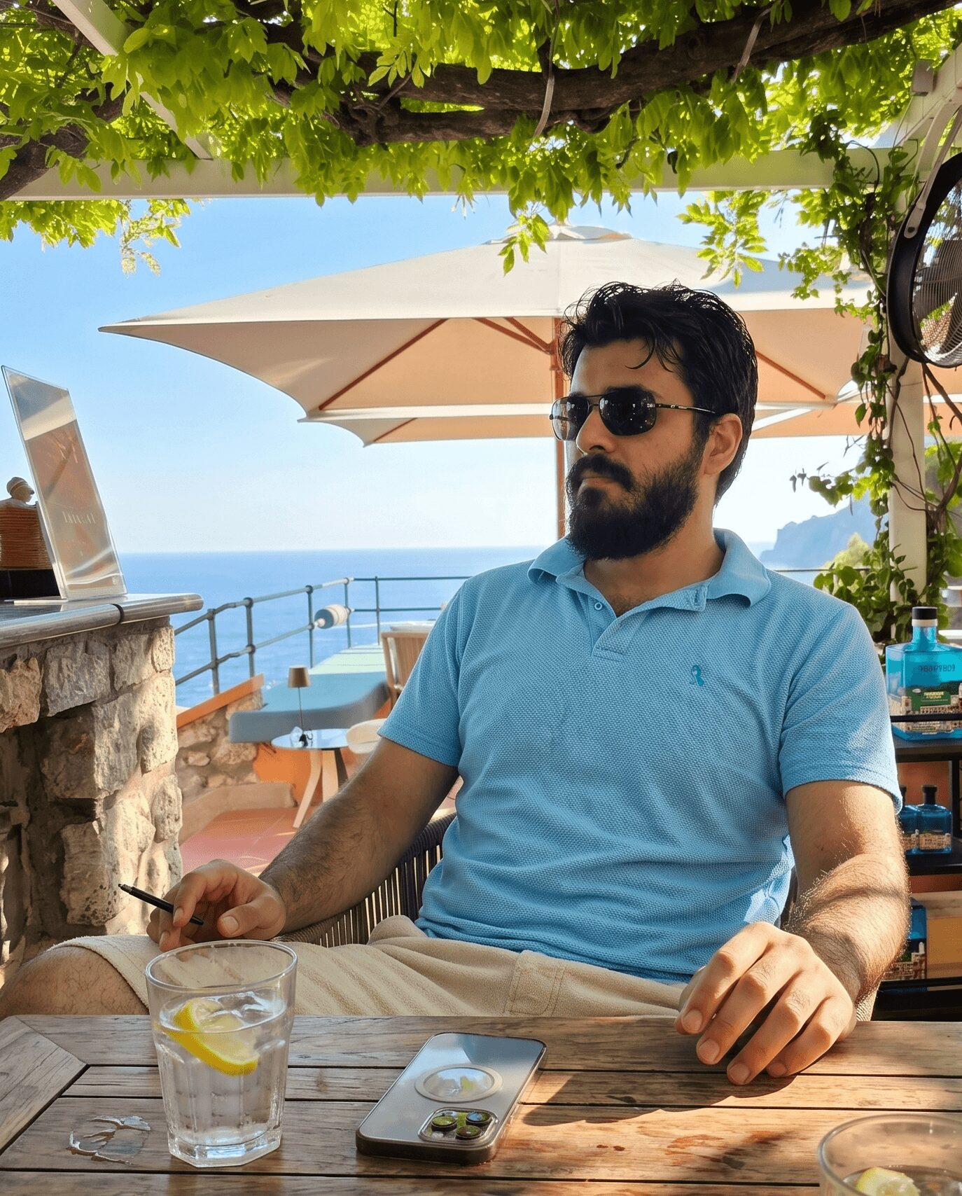 Man with dark hair and beard wearing sunglasses and a blue polo shirt sitting outdoors at a table with a view of the sea, under a canopy with green leaves.