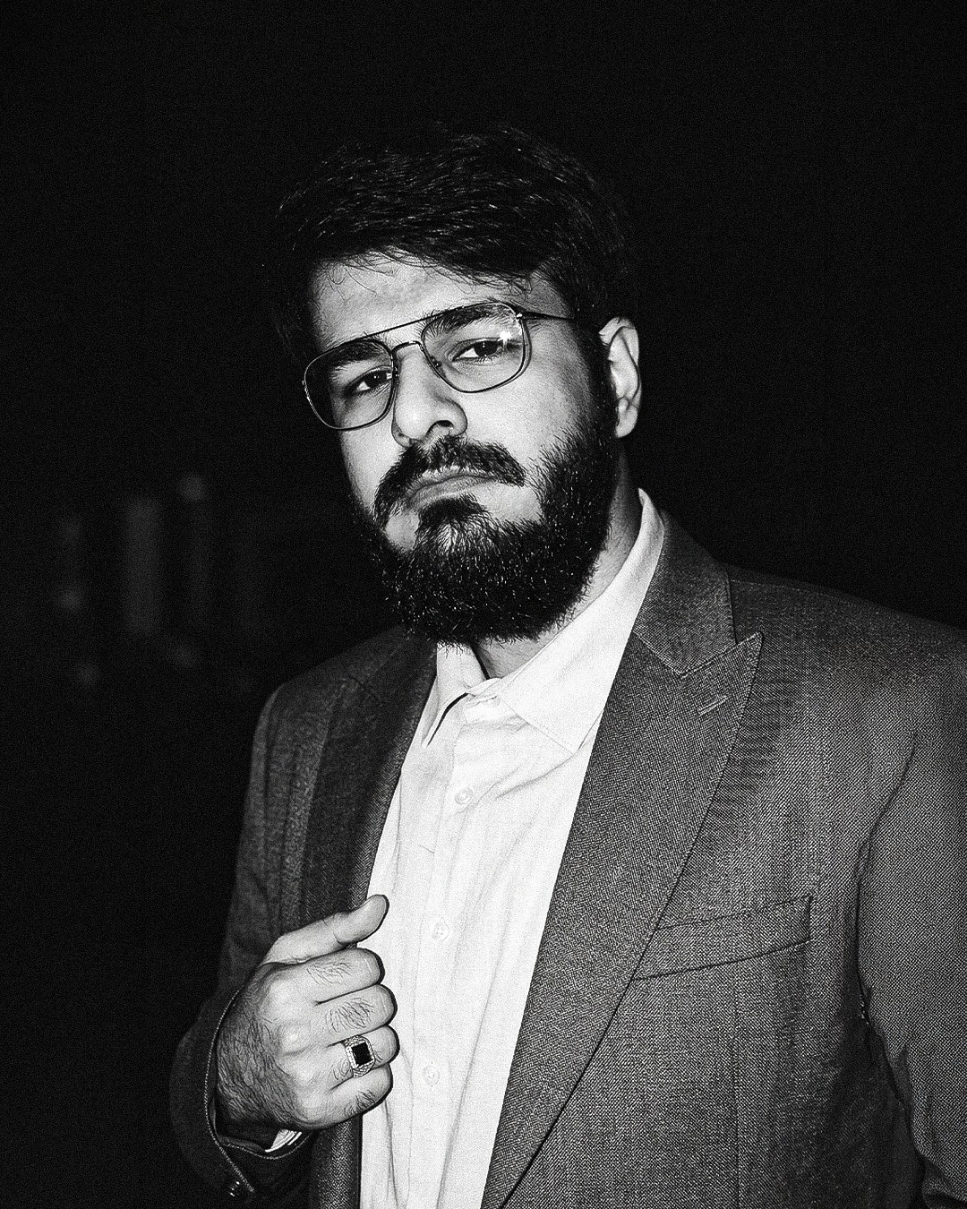 Man with glasses, beard, and mustache in a suit, adjusting his jacket, looking serious, in black and white.