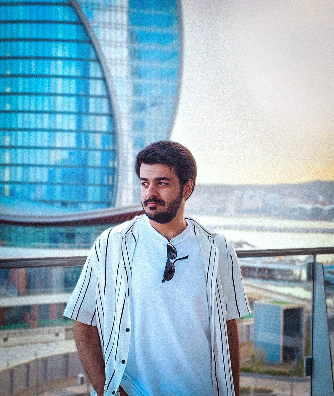 Young man with dark hair and beard stands outdoors near a modern glass building, wearing a light blue shirt and sunglasses hanging from collar.