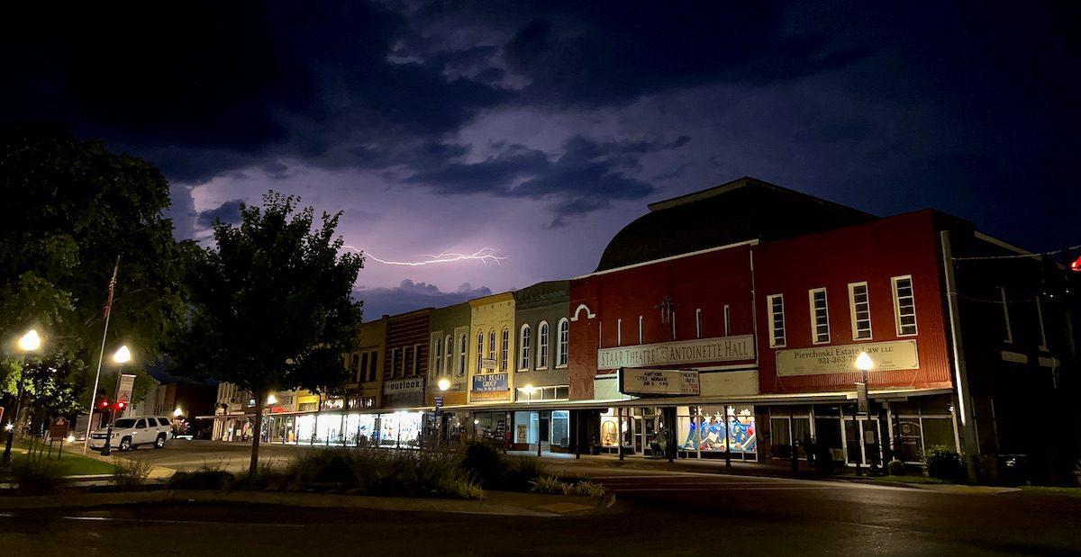 Night scene of a colorful building with trees and streetlights, lightning in the dark cloudy sky, and a parking lot.