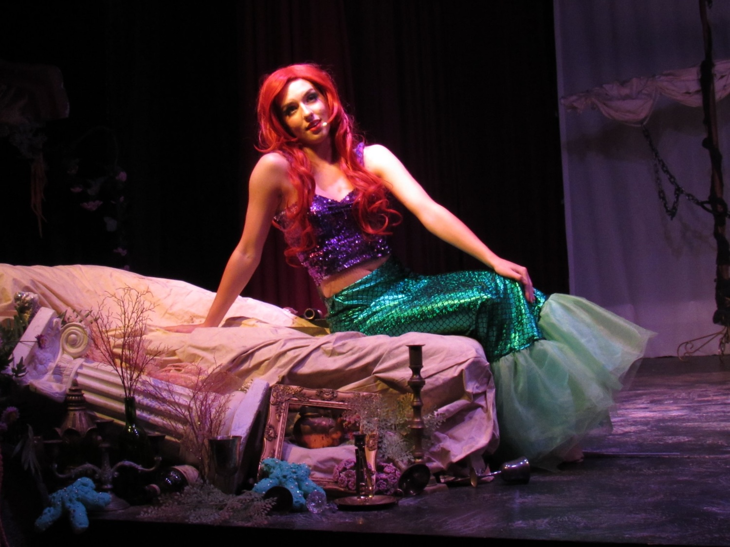 Woman in a colorful costume sitting on a bed on stage, with a dark background and stage props around.
