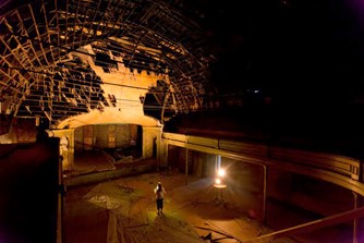 Person standing in an empty, dark theater with a damaged ceiling and a bright light on the stage.