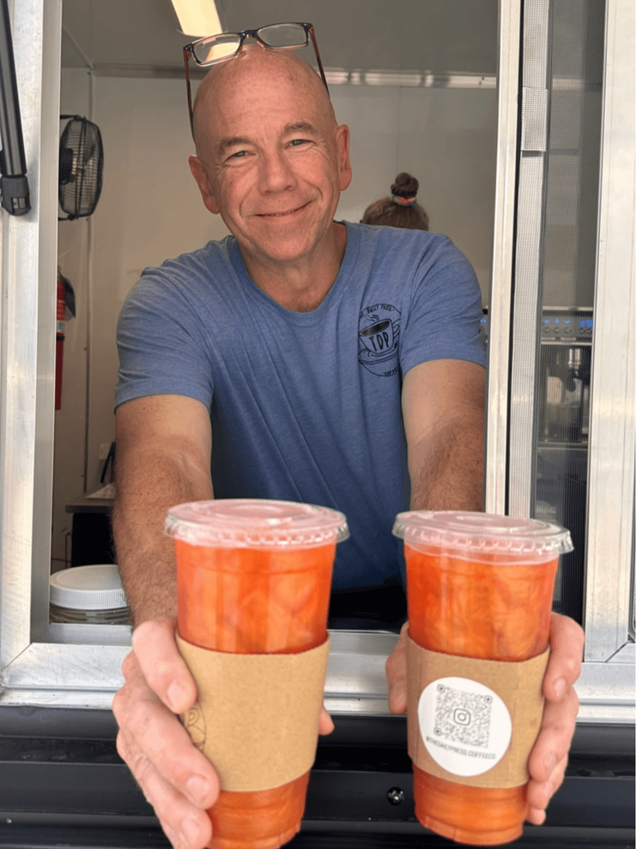 Person with glasses on head holding two cups of iced tea or coffee, smiling, in a cafe setting.