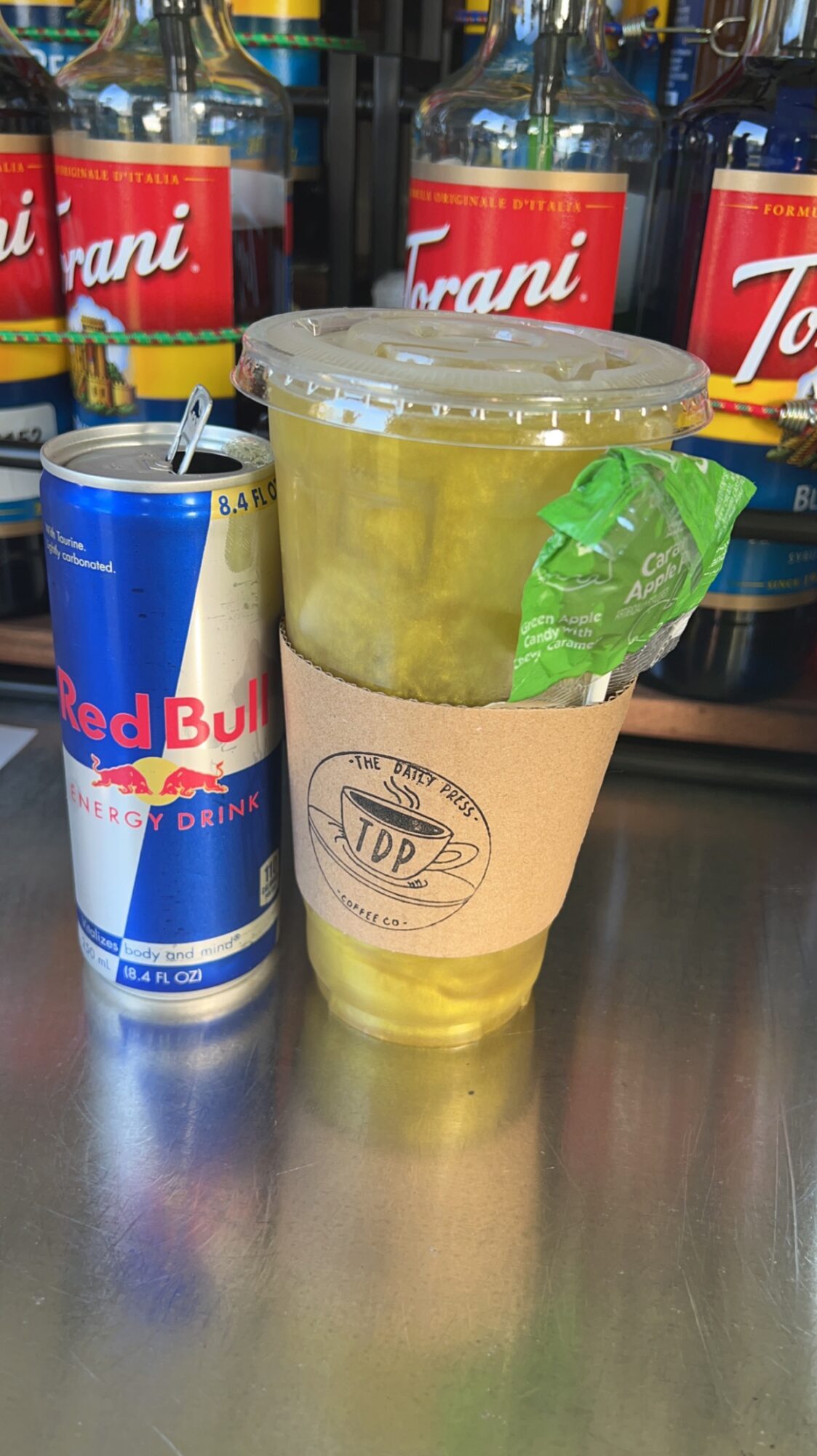 Plastic cup with yellow drink and ice, straw, and a green packet, next to a Red Bull can, on a reflective surface.