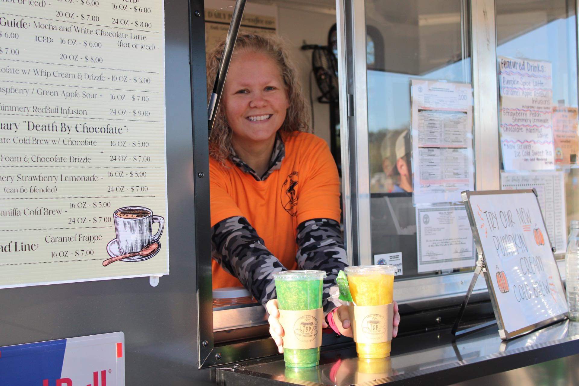 Smiling person in orange shirt behind a counter with colorful cups, menu, and glass window, in a food stand or cafe.
