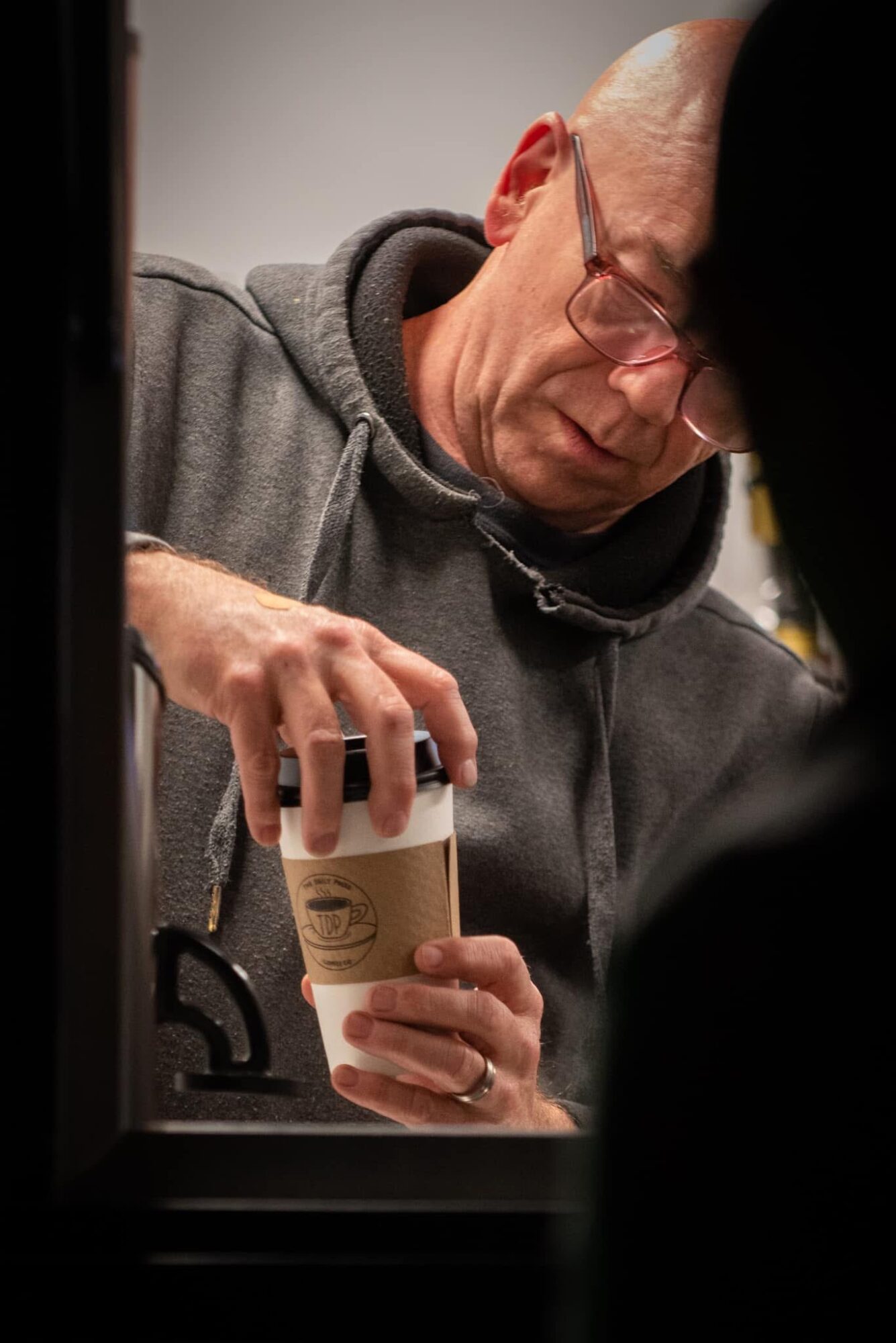 Man with glasses in a hoodie holding a coffee cup, viewed through a window or opening.