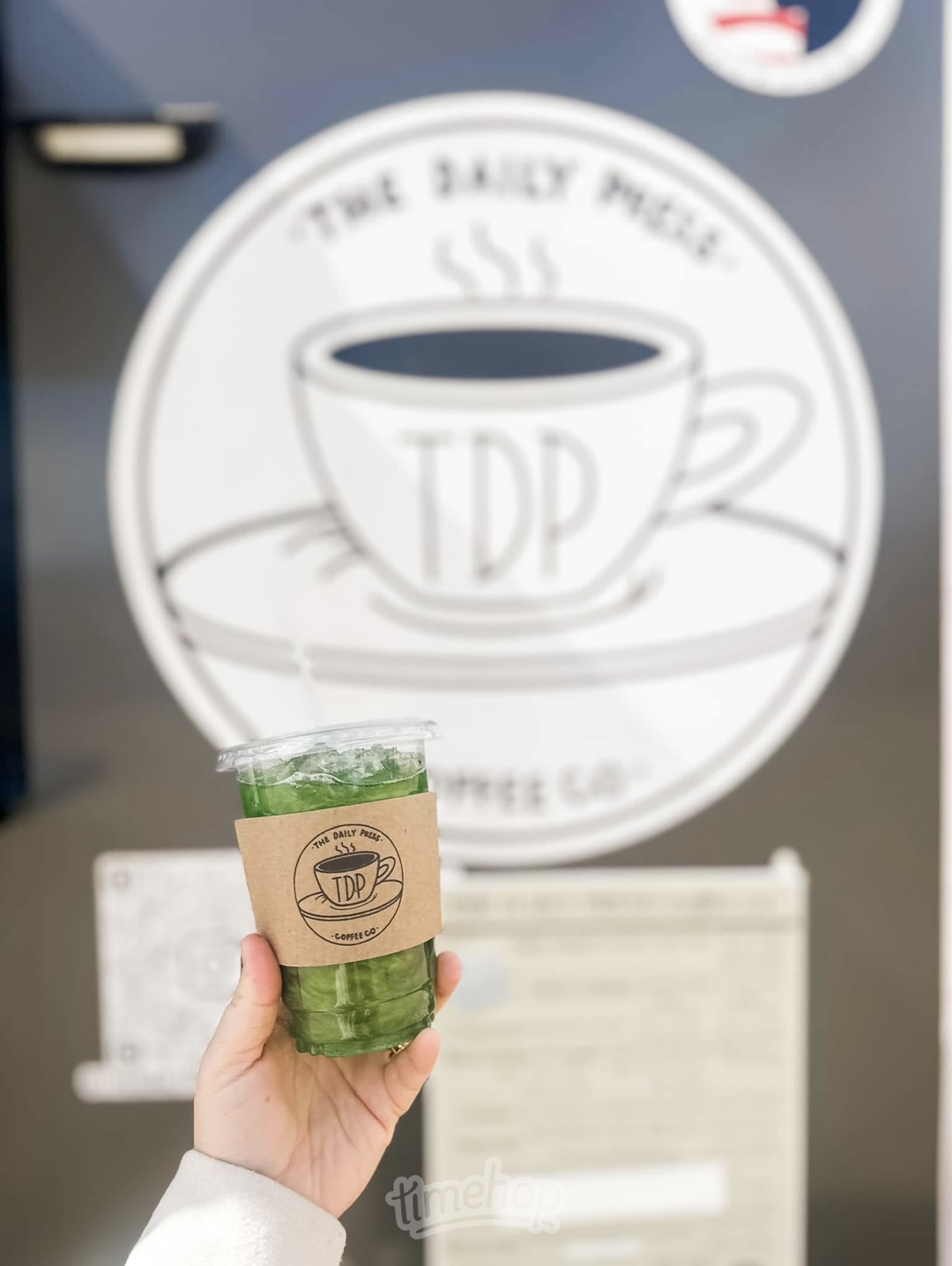 Hand holding a cup of green tea with a brown sleeve, in front of a circular sign with a coffee cup illustration and text.