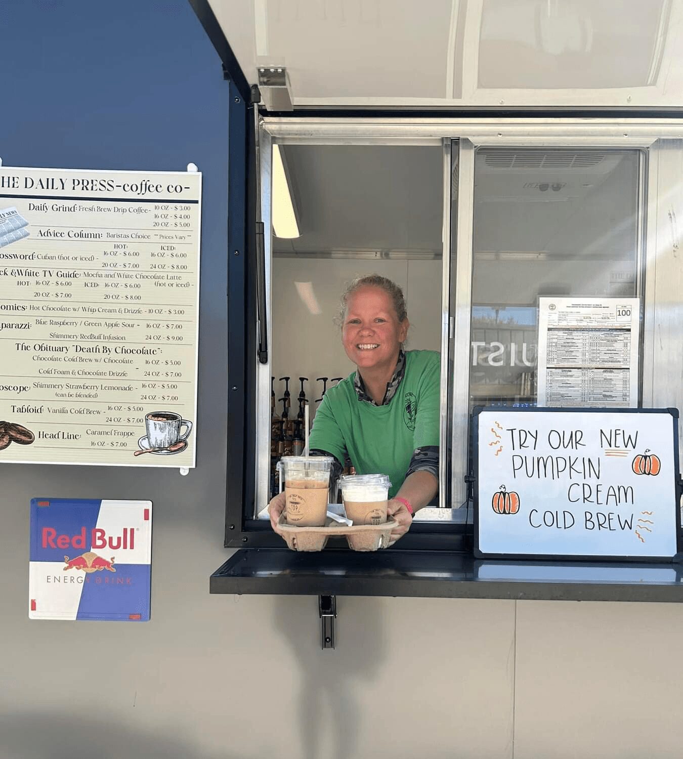 Smiling woman in green shirt behind a window serving pumpkin ice cream and cold brew coffee, with signs and posters nearby.