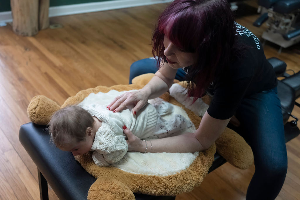 Adult woman leaning over a baby lying on a cushioned changing pad on a wooden floor, in a room with exercise equipment.