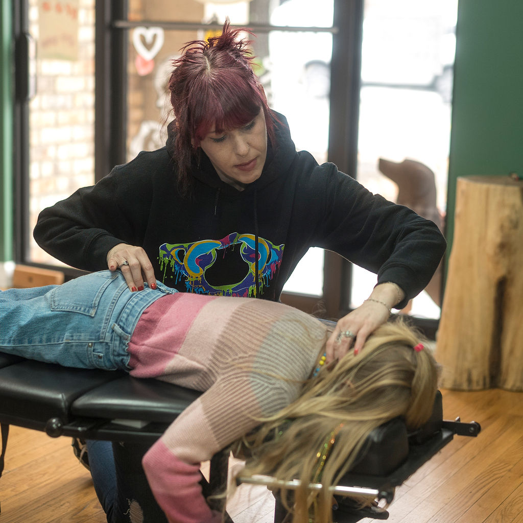 A woman with red hair performs a massage on a person lying face down on a massage table inside a room with large windows and wooden floors.