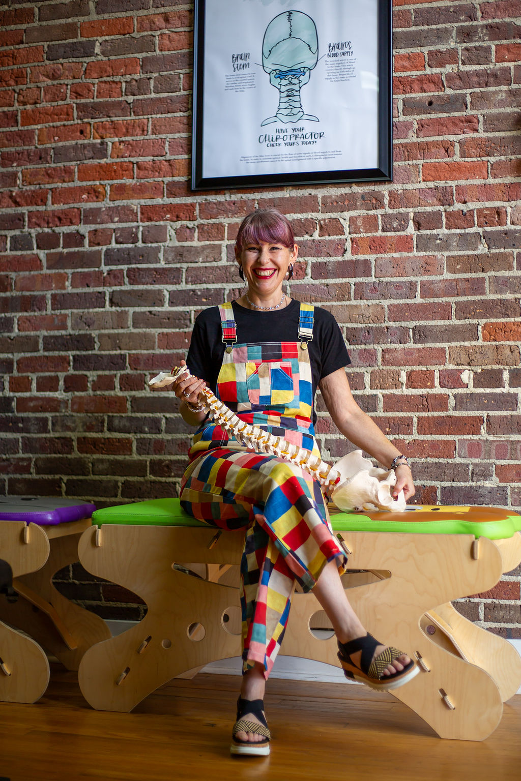 Woman with short hair and colorful checkered pants sitting on a wooden bench, holding a dog, against a brick wall with framed artwork.