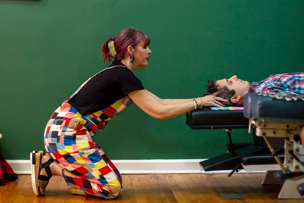 Woman kneeling and reaching out to a person lying on a medical table, with a green wall in the background.