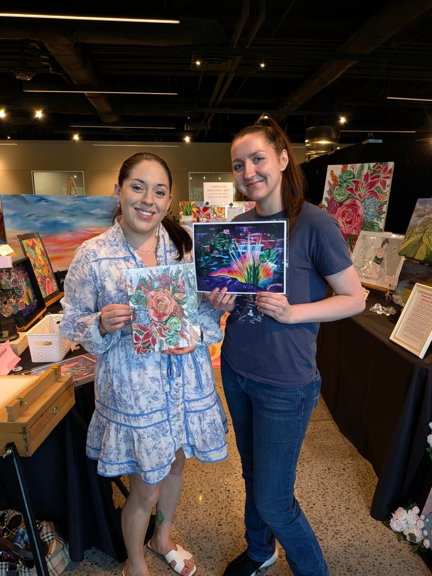 Two women holding colorful paintings in an art gallery, smiling at the camera.