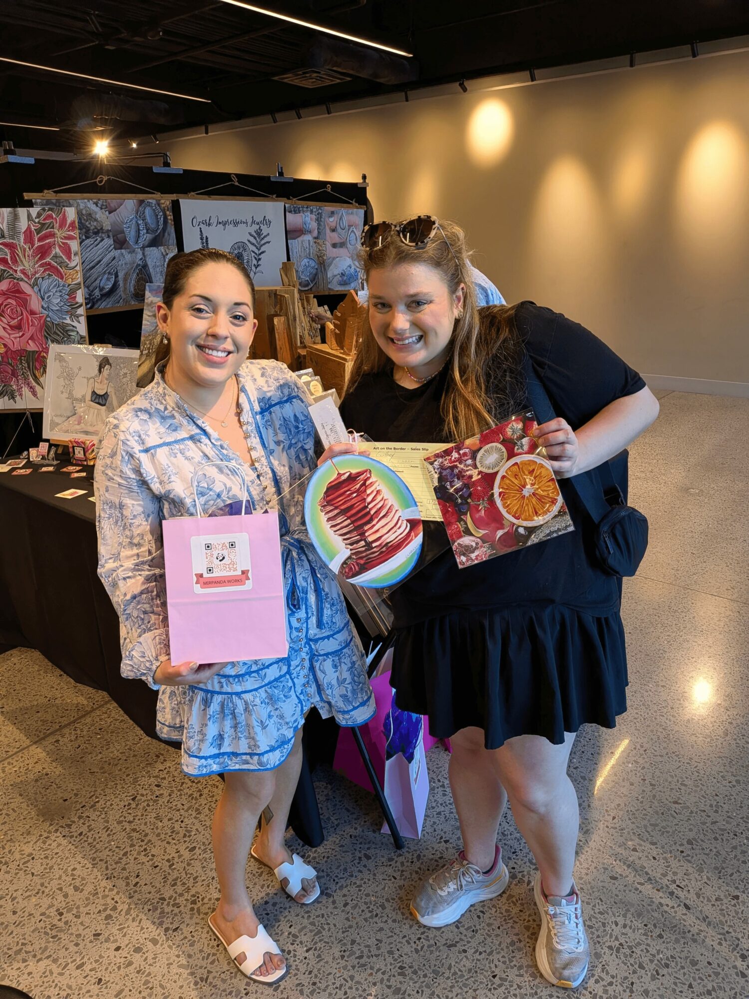 Two smiling women holding colorful artwork in an indoor market or exhibition space.