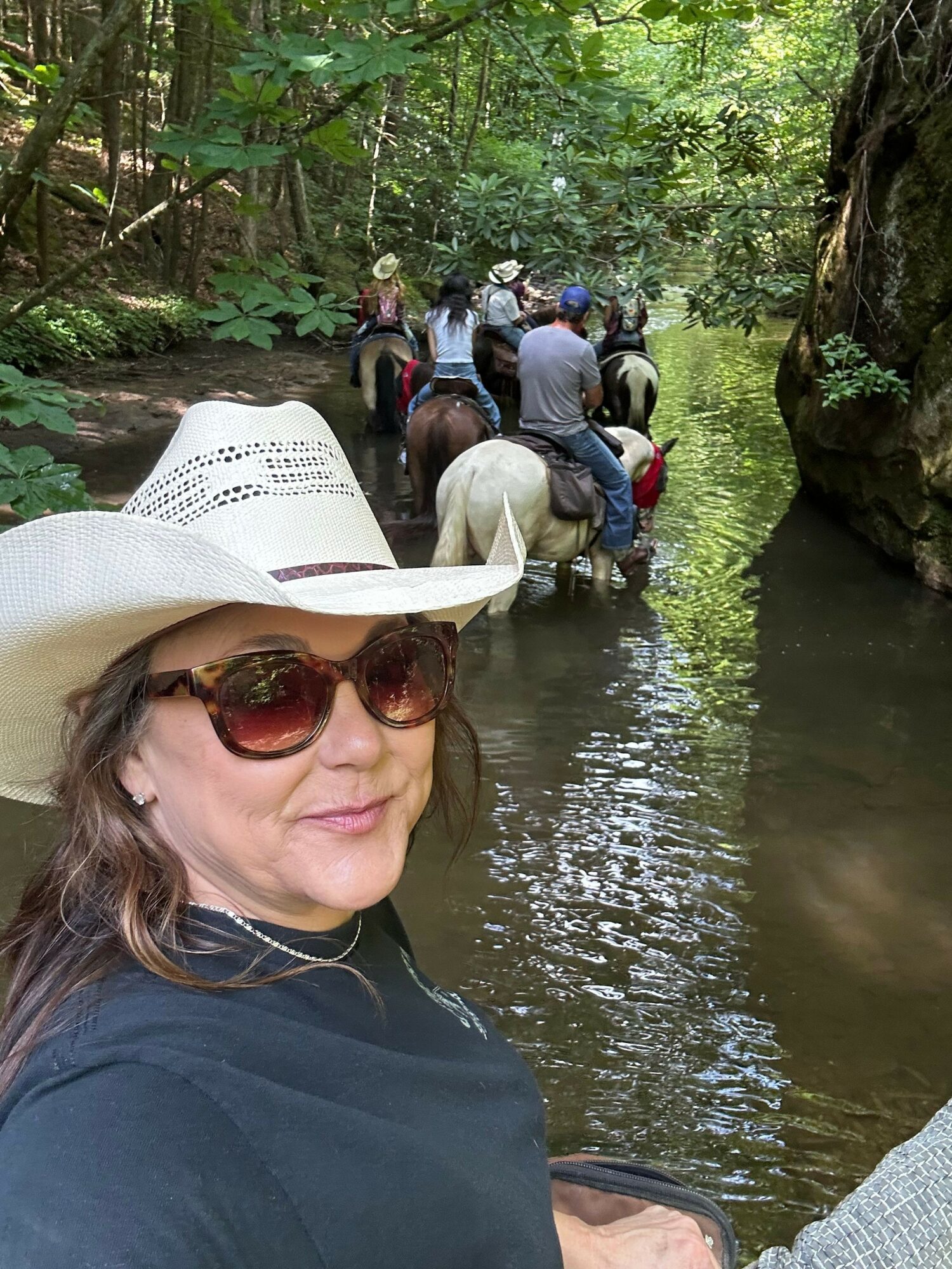 Woman wearing sunglasses and a white hat taking a selfie near a river with a group of people in the background.