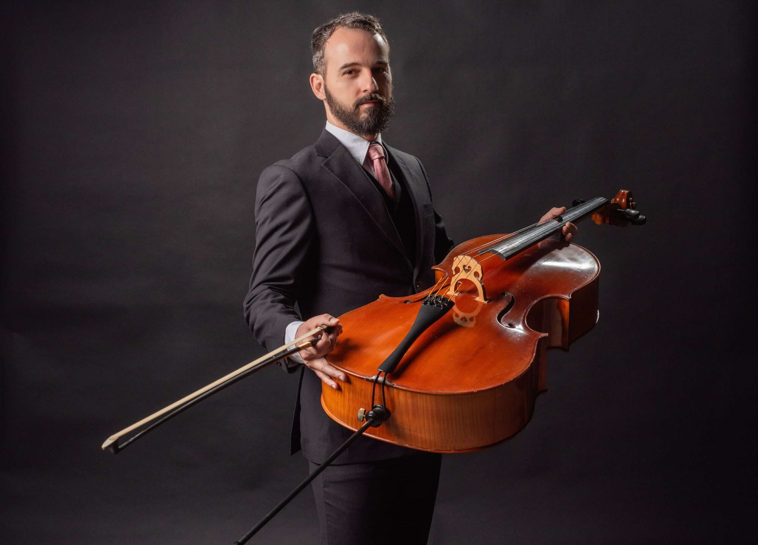 Man in suit holding a cello and bow against a dark background.