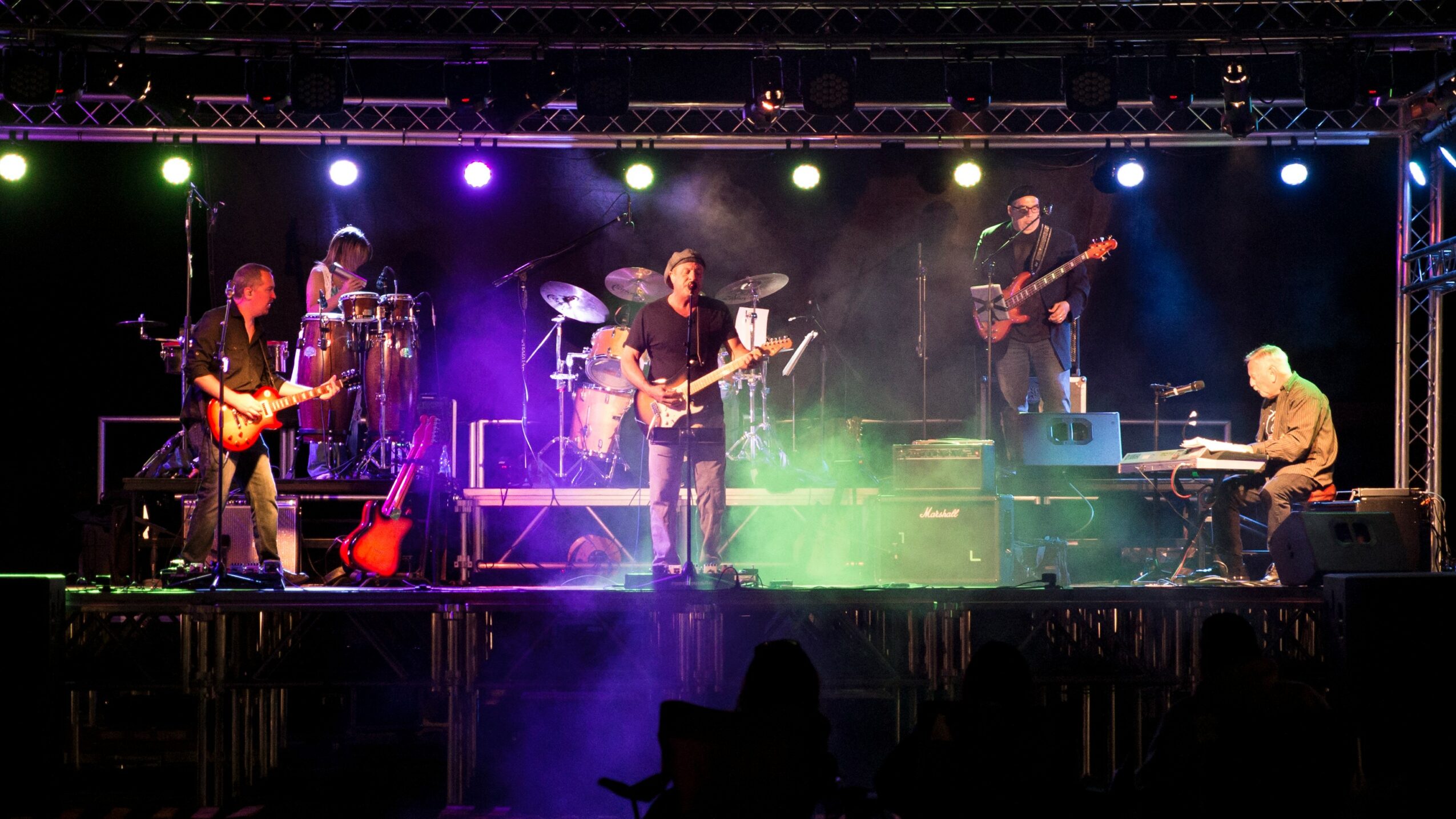 Band performing on stage with colorful lights, musicians playing guitars, keyboard, and drums, and fog effects.
